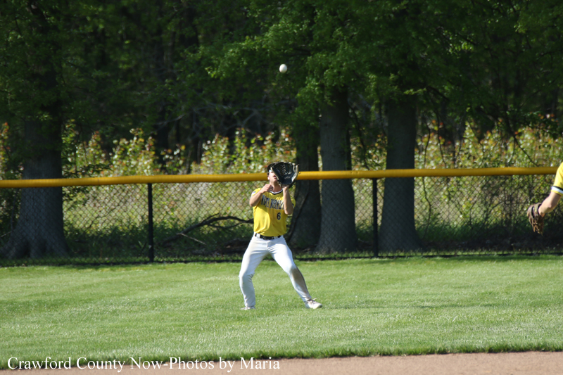 Young baseball player in a yellow uniform catching a ball with his glove in a grassy field near a fence.