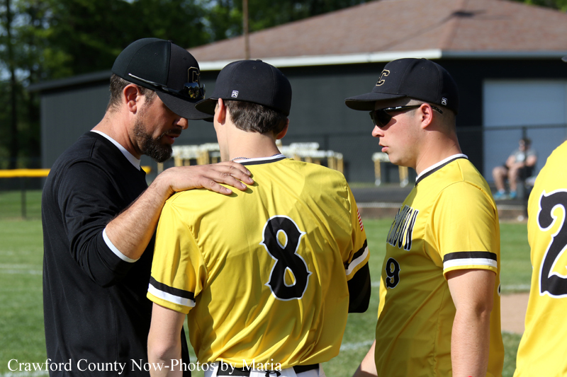 Baseball coach talks to two players in yellow uniforms on a sunny field, one with an arm on the other's shoulder.