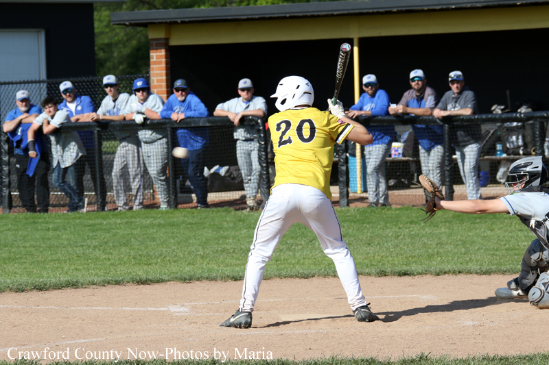 Baseball batter in yellow jersey #20 swings at a pitch near the catcher and home plate as spectators watch from the dugout fence behind him.