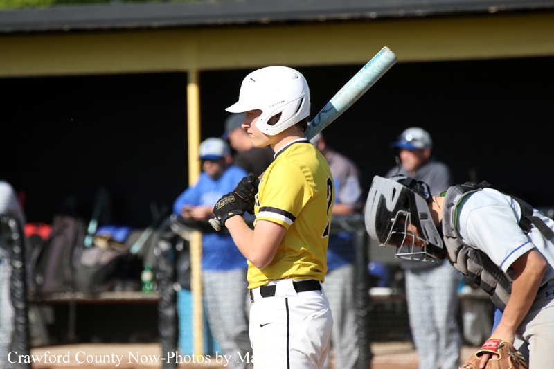 Baseball batter in a yellow uniform and white helmet stands at the plate, bat resting on his shoulder; catcher in gray gear crouches behind him.