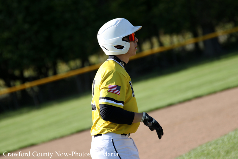 Baseball player in a yellow jersey and white helmet jogging on a sunny baseball field, with red sunglasses and a black undershirt.