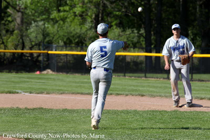 Baseball pitcher wearing #5 mid-throw on a sunny field while another fielder watches from the infield with a ball in the air.