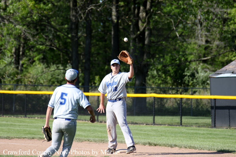 Two baseball players in gray uniforms on a dirt infield; one throws the ball while the other watches, with a yellow fence and trees in the background.