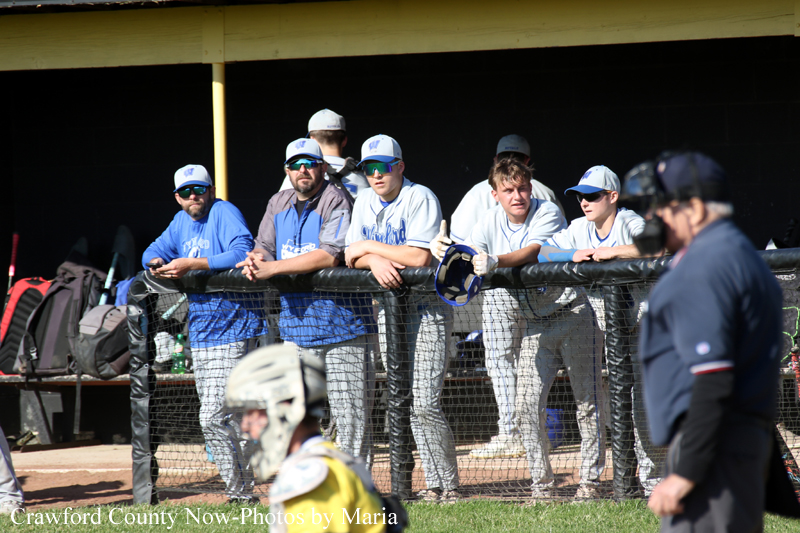 Group of baseball players leaning on the dugout railing and watching a game, some wearing blue and gray uniforms and caps.