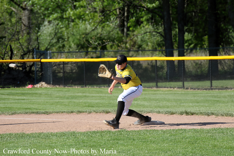A baseball player in a yellow jersey and black cap fielding a ground ball, glove extended, on the infield dirt with a grassy outfield behind him.