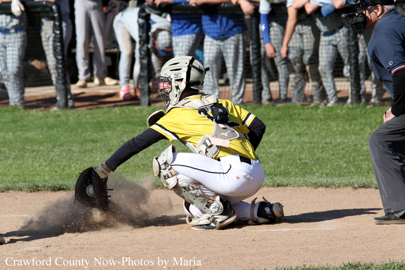 Baseball catcher in yellow and white gear sliding into home plate as dust rises, with spectators in the background.