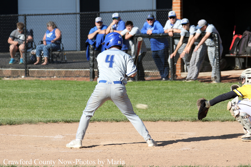 Youth baseball batter in gray uniform and blue helmet (number 4) at home plate preparing to swing; catcher in yellow crouches nearby as crowd watches behind the fence.