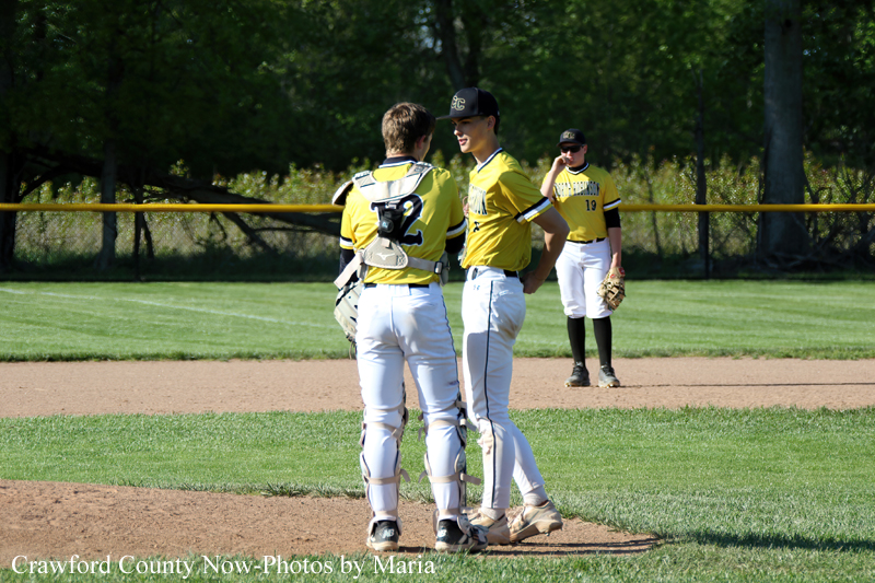 Two yellow-uniform baseball players chat on the infield; one wears catcher gear while the other stands with hands on hips, with a fielder in the background.