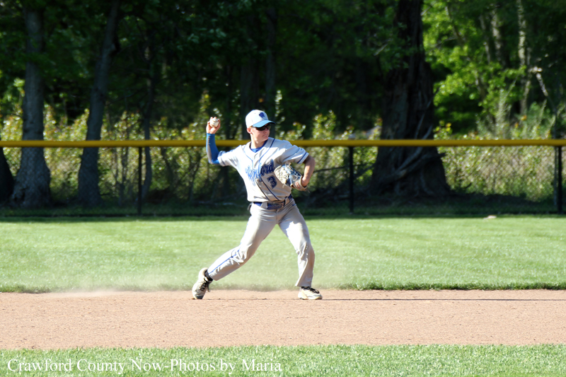 Right-handed baseball pitcher in gray uniform delivering a pitch on a dirt infield, with grass and trees in the background.