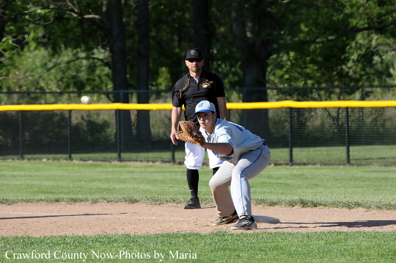 Baseball player in a gray uniform crouches at infield with glove as a ball approaches; an umpire stands behind on a sunny field.