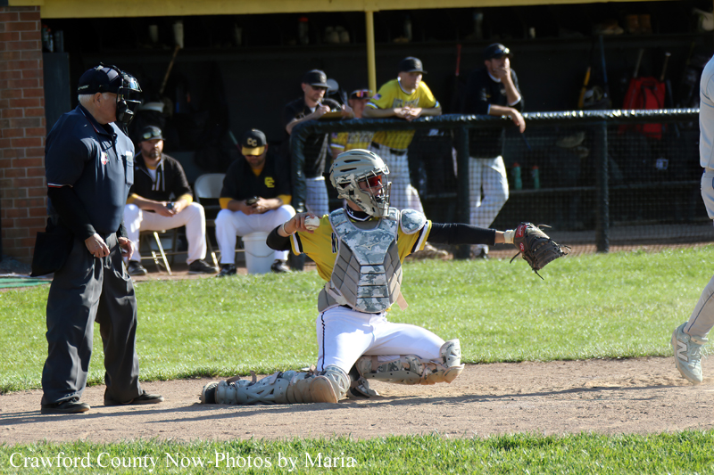 A baseball catcher slides on one knee at home plate with glove extended, while an umpire watches and teammates sit in the dugout in the background.