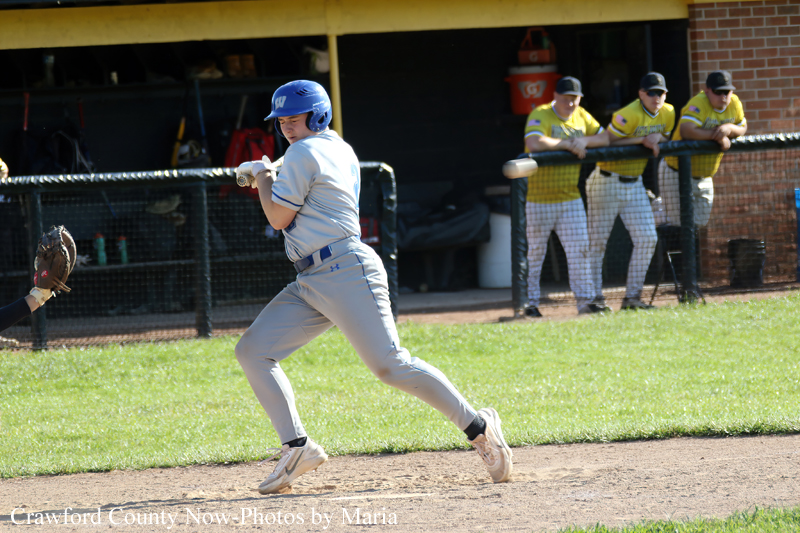 Baseball player in a gray uniform and blue helmet runs toward first base with a bat in hand, dugout with teammates in yellow jerseys watching in the background.