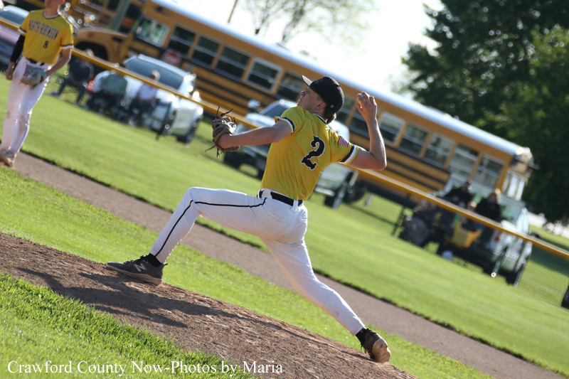 Baseball pitcher in a yellow jersey winds up to throw a pitch on the mound with teammates and a school bus in the background.