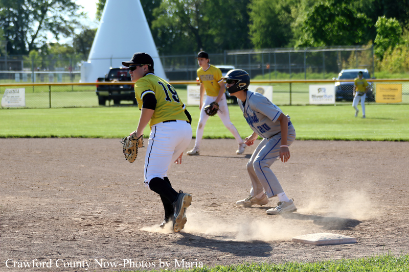 Two youth baseball players in a game: a gray-uniform baserunner near second base while a yellow-uniform fielder moves in with a glove to tag, dust rising from the dirt.