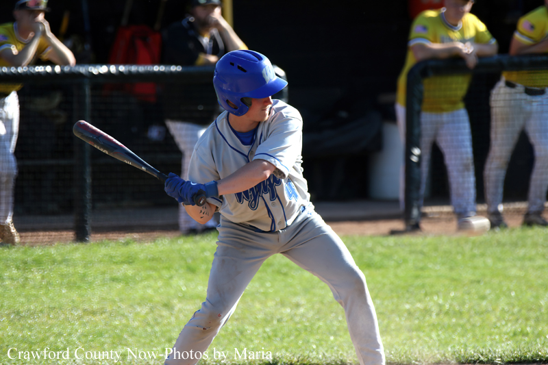 Youth baseball player in a gray uniform and blue helmet swings a bat mid-at-bat on a sunny field.