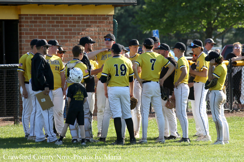 Youth baseball team in yellow jerseys huddle with coach on a sunny field, players wearing numbered uniforms.