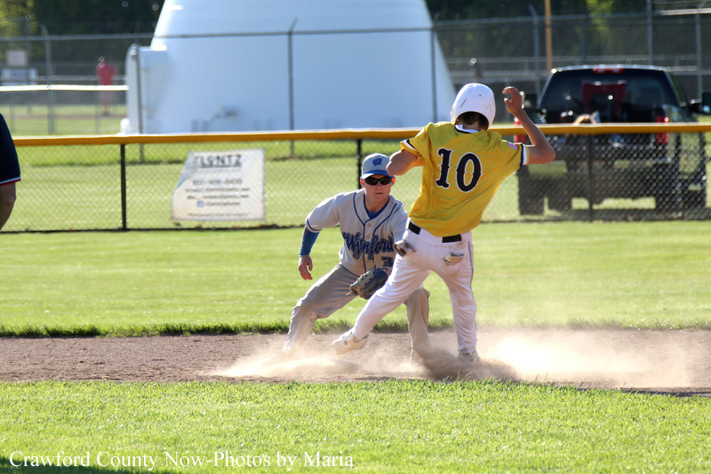 Two young baseball players on a sunny field: a yellow-uniform runner (#10) slides into a base while a gray-uniform fielder pursues, kicking up dust.