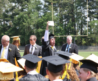 Graduate in cap and gown raises diploma above head in a loud celebration on a football field, surrounded by fellow graduates and faculty attendees.