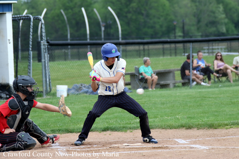 Young baseball batter in a white uniform and blue helmet swings at a pitched ball, with the catcher crouched behind him on the dirt field.