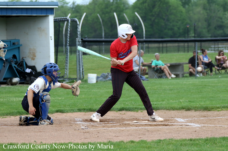 Youth baseball game: batter in a red shirt and white helmet swings at a pitch while a blue-gear catcher crouches behind home plate; spectators watch beyond the fence.