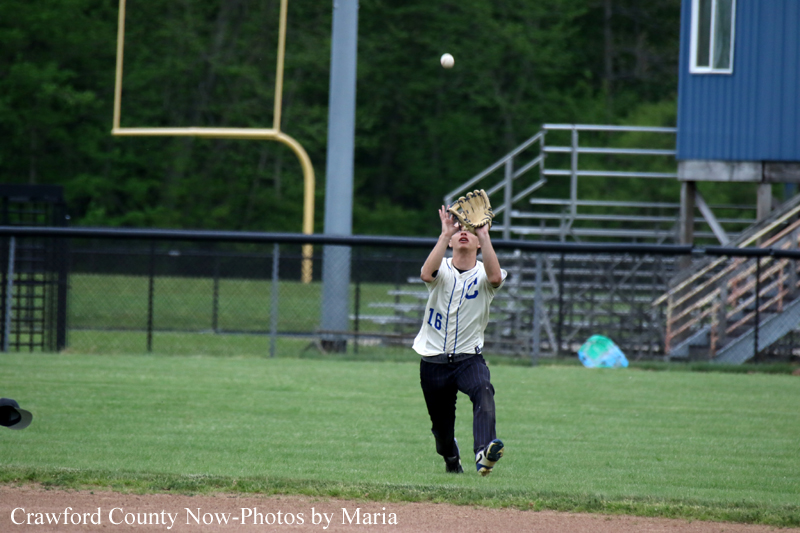 Young baseball player in a white jersey (number 16) reaches up with a glove to catch a high fly ball on a grassy field near the outfield fence.