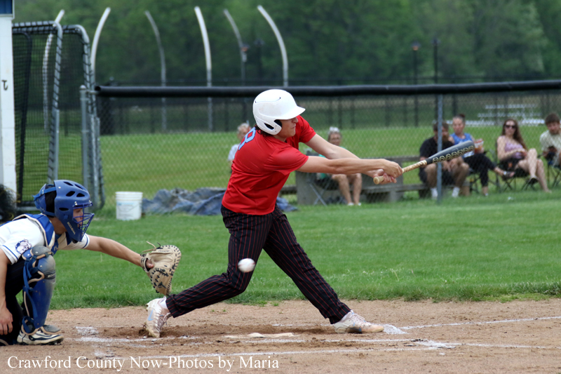 Baseball batter in red jersey swings at a pitch while the catcher in blue gear crouches behind home plate and spectators watch from the fence.