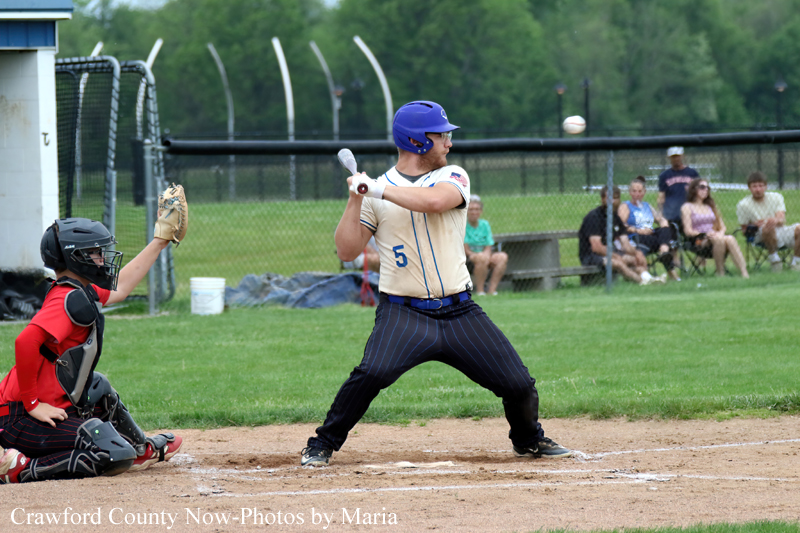 Baseball batter in a tan uniform and blue helmet swings at a pitch as the catcher in red protective gear crouches behind home plate.