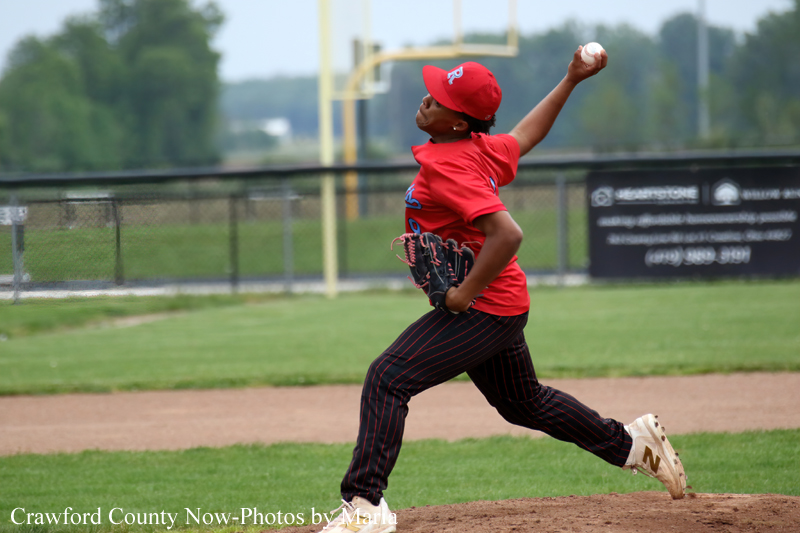 Young baseball pitcher in a red uniform winding up to throw on a sunny field as a ball leaves his hand
