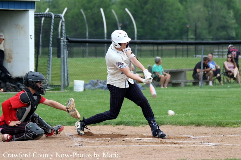 A batter swings at a pitch near home plate as a catcher crouches behind, with a green field and fence in the background.