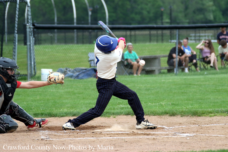 Young baseball batter in a white jersey and blue helmet swings at a pitch at home plate, with a catcher reaching and spectators in the background.