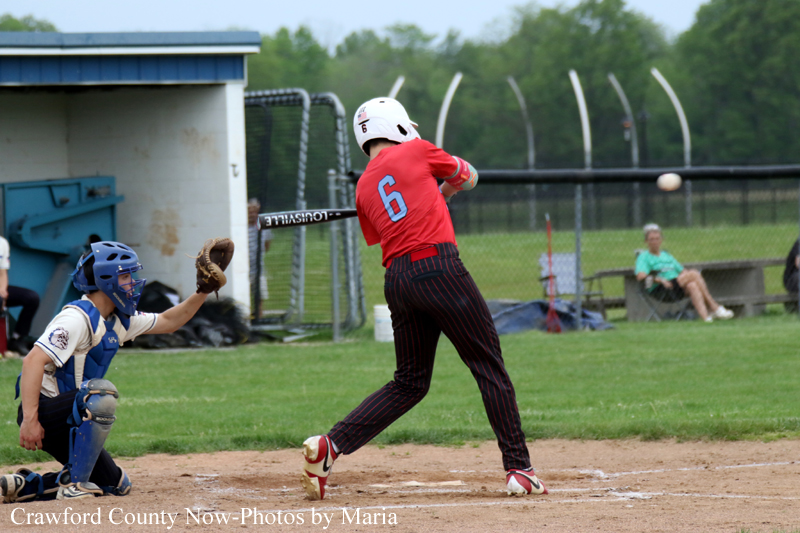 Baseball batter in a red jersey with number 6 swings at a pitch as the catcher crouches behind home plate on a grassy field.