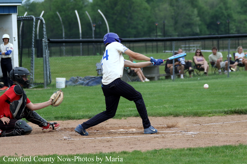 Baseball batter in white jersey swinging at a pitch as the catcher in red and black gear crouches behind home plate.
