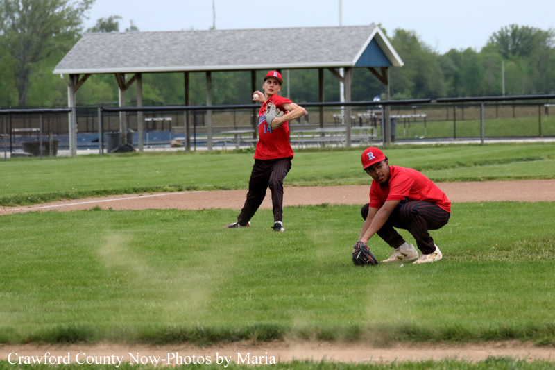 Two baseball players on a grassy field: a pitcher in red throws a ball while a fielder in red crouches nearby by the infield dirt.