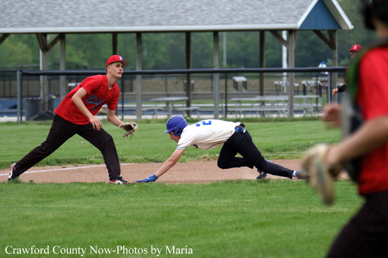 Baseball baserunner in white and blue slides headfirst toward a bag as a fielder in red stands nearby on the infield dirt.