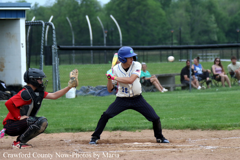 Youth baseball batter in white jersey and blue helmet swings at a pitch, with catcher in red/black gear crouched behind home plate on the dirt field.