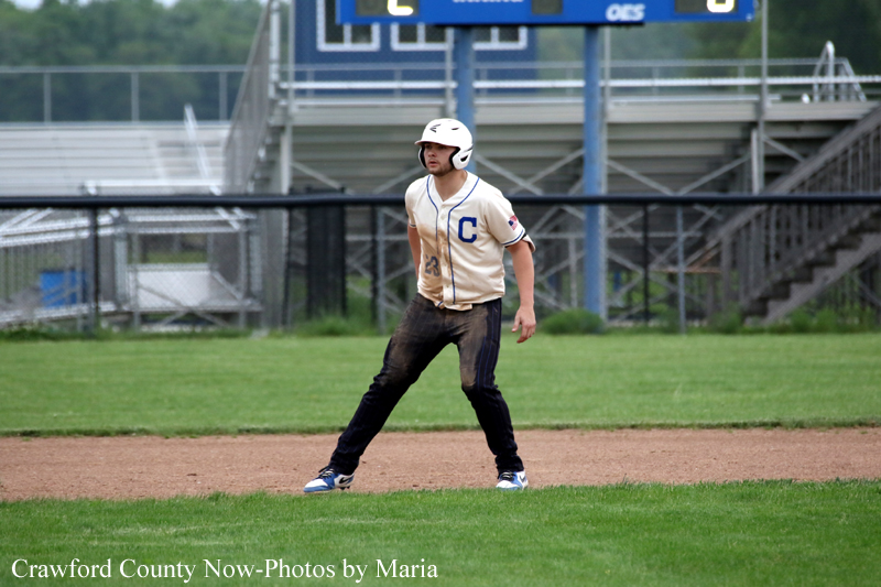 Baseball player in a beige jersey with a blue 'C' stands on infield dirt, helmet on, ready to play with the scoreboard in the background.