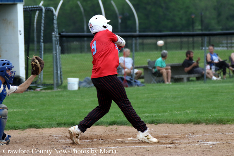 Baseball batter in a red jersey and white helmet swings at a pitch as the catcher in blue crouches behind him on the dirt infield with a green outfield and spectators in the background.