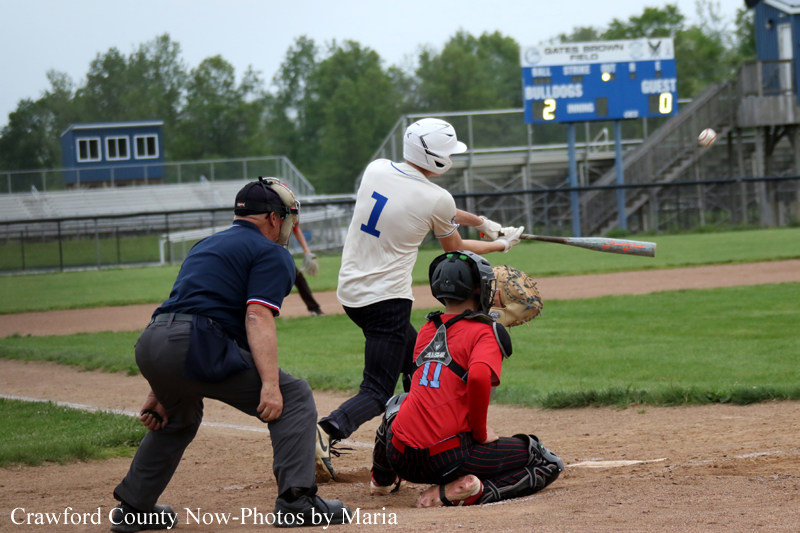 Baseball batter in white swings at a pitch as catcher in red kneels behind home plate; umpire stands nearby on a dusty field with a scoreboard in the background.