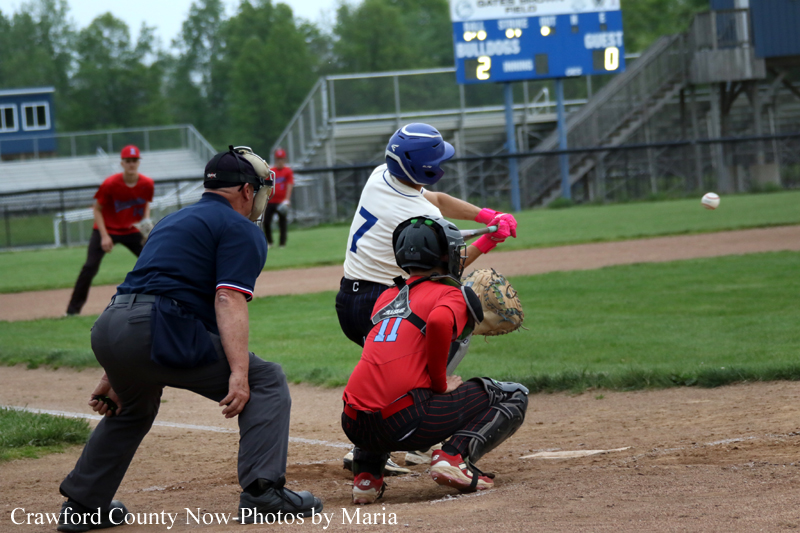Youth baseball: batter in white and blue swings as catcher in red crouches behind home plate with umpire nearby.
