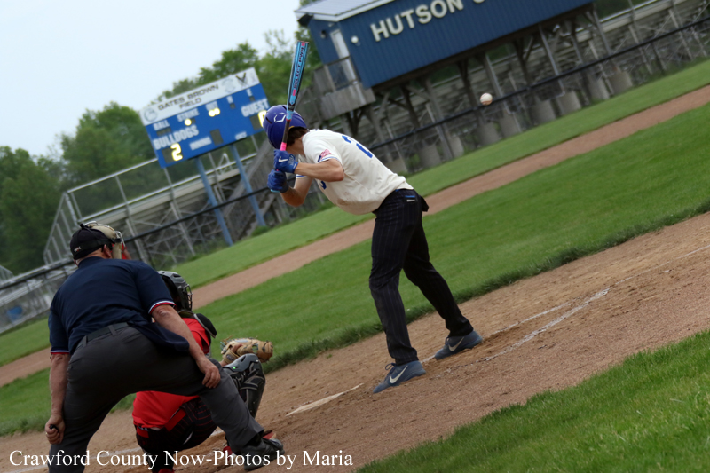 Baseball batter in a white uniform and blue helmet stands at home plate, ready to swing, with catcher and umpire crouched behind him on the dirt and a scoreboard and blue grandstand in the background.
