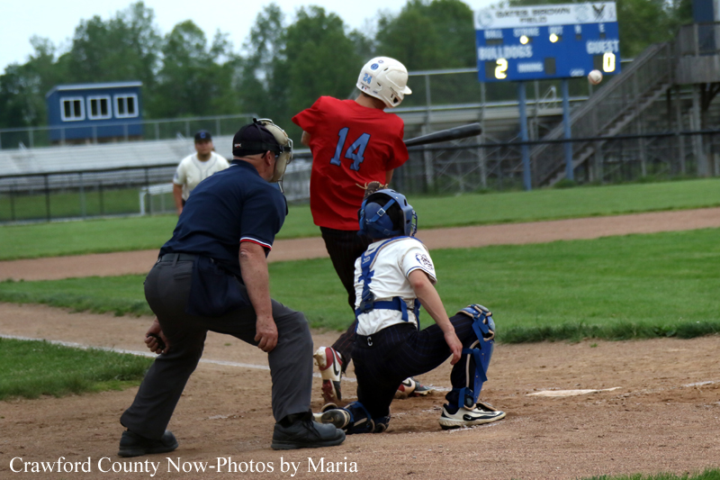 Young batter in red jersey swings at a pitch as the catcher and umpire crouch behind home plate on a sunny baseball field.