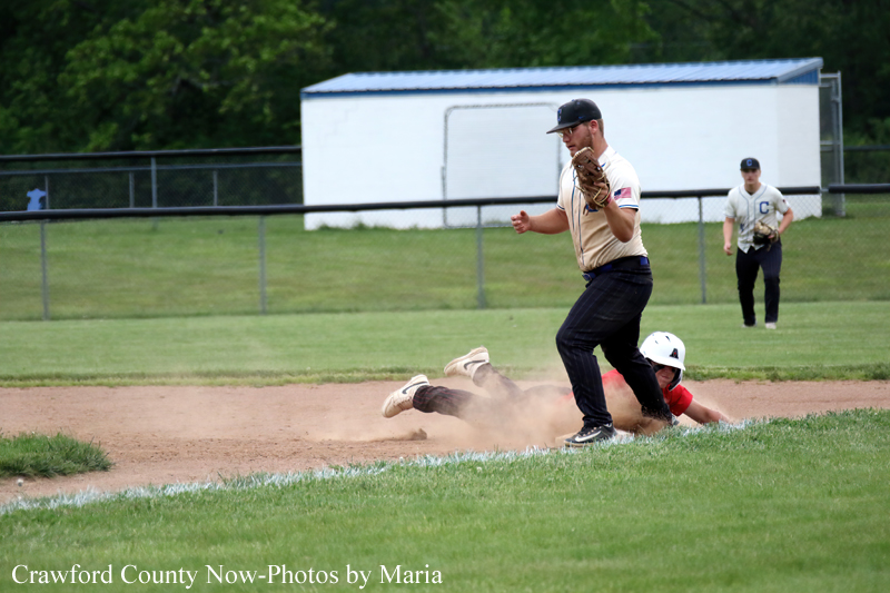 Baseball: a runner in a tan uniform slides into a base, kicking up a dust cloud while a fielder watches from the infield.