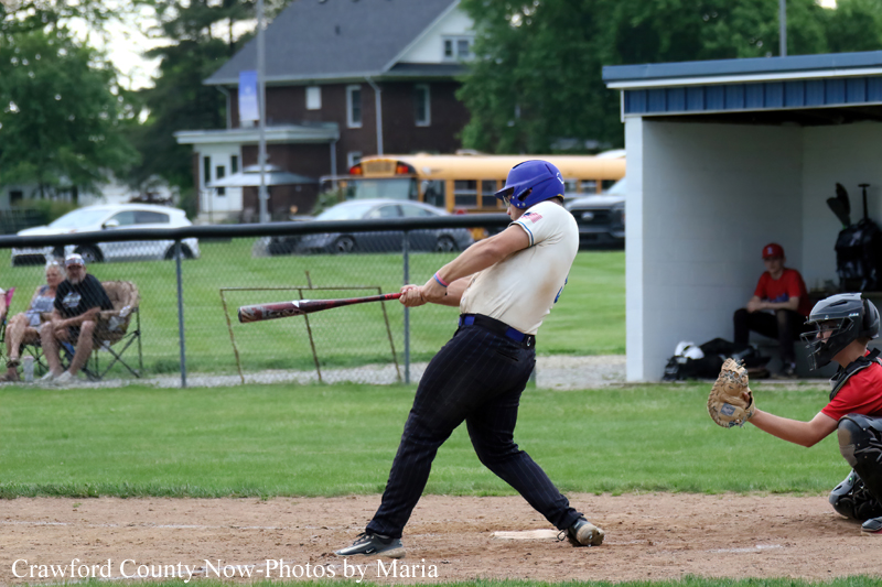 Baseball batter in white jersey and blue helmet swings at a pitch at home plate as the catcher crouches behind him on a dirt field.