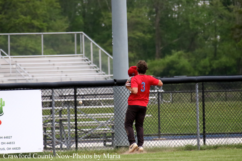 Baseball player in a red jersey #3 leans on the outfield fence during a game.