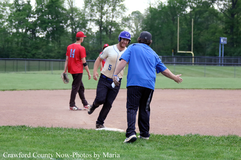 Baseball baserunner in a white jersey with number 5 slides toward a base as a coach in a blue shirt extends his arms to greet him on the dirt infield.