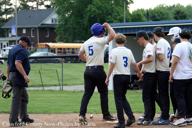 Baseball coach on the left tipping his helmet as players in beige uniforms with blue numbers line up at home plate.