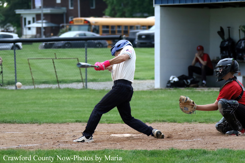 Baseball batter in white jersey swings at a pitch, with a catcher in red and black crouched behind home plate.