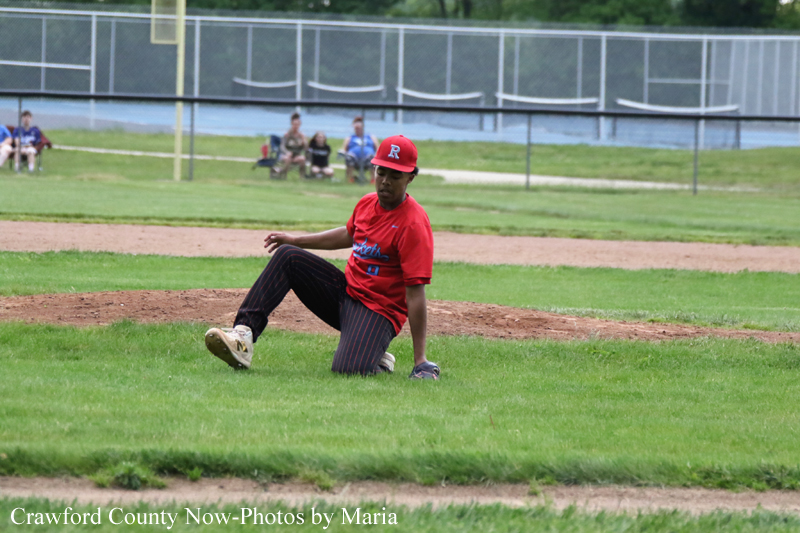 Young baseball player in a red uniform kneels on the pitcher's mound during a game, with a dirt infield and green grass surrounding him.