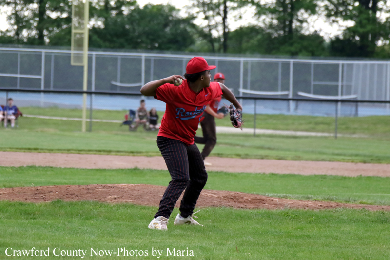 Young baseball pitcher in a red uniform on the mound, mid-throw during a game on a grassy field.