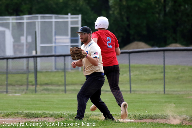 Baseball players in a field: a beige-uniform fielder with a glove and cap watches a play as a runner in red (#2) with a helmet passes by behind him on the grass.